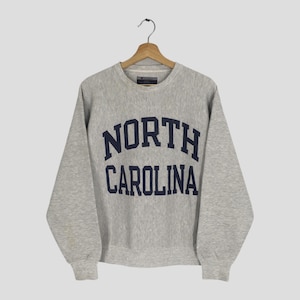 May include: A heather gray sweatshirt with the words "NORTH CAROLINA" in navy blue block letters. The crew neck sweatshirt is hanging on a wooden hanger.