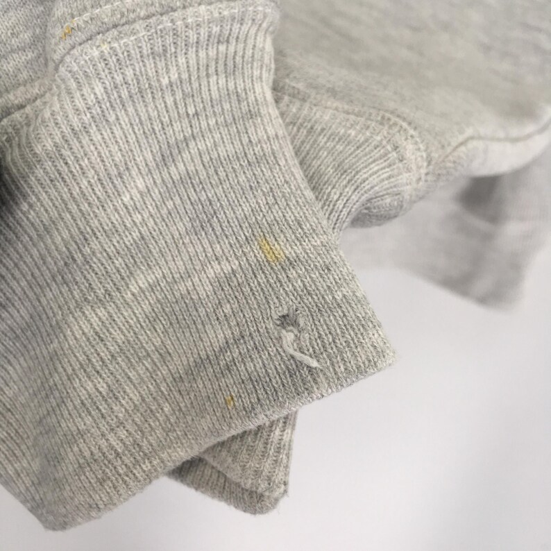 May include: Close-up of a light gray sweatshirt sleeve. The ribbed cuff shows wear and tear, with a small hole and several yellow stains. The fabric appears soft and slightly worn.