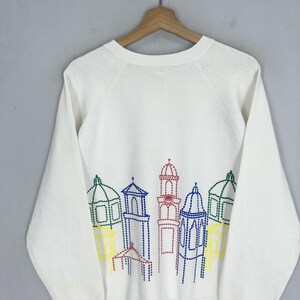 May include: White long-sleeve sweatshirt featuring a colorful embroidered cityscape design across the chest. The design includes various buildings outlined in blue, green, red, and yellow thread. The sweatshirt is hanging on a wooden hanger.