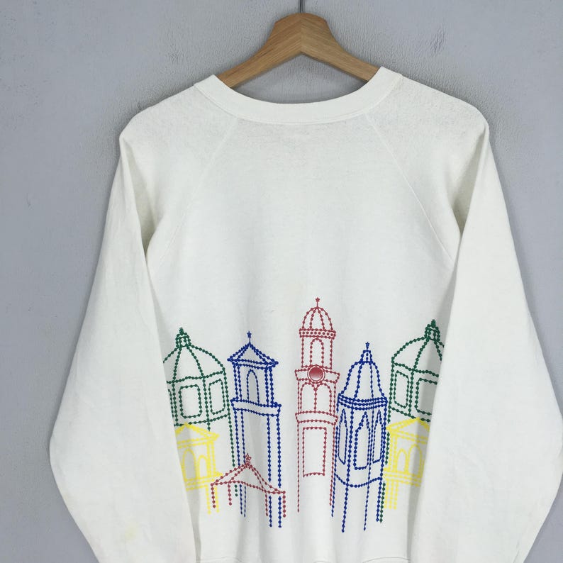May include: White long-sleeve sweatshirt featuring a colorful embroidered cityscape design across the chest. The design includes various buildings outlined in blue, green, red, and yellow thread. The sweatshirt is hanging on a wooden hanger.