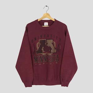 May include: A maroon crewneck sweatshirt with the words "UNIVERSITY OF MINNESOTA GOPHERS" printed on the front. The graphic includes a large "M" logo in brown and gold, with a laurel wreath design. The sweatshirt is hanging on a wooden hanger.