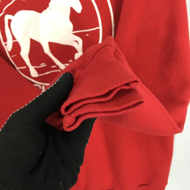 May include: A red sweatshirt with a white horse graphic inside a circle. The sleeve cuff is folded, revealing the inside of the red fabric. The sweatshirt is made of a soft material.