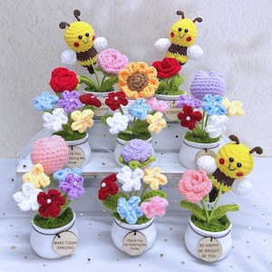 May include: A collection of handmade crochet flowers in small white pots. The flowers are various colors, including red, yellow, pink, and blue. Some pots have small wooden tags with phrases like "Make Today Amazing". Two pots have yellow and brown crochet bees.