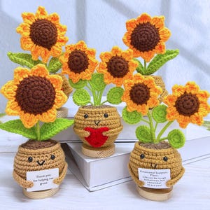 Op de afbeelding: Gehaakte zonnebloemen met bruine centra en gele bloemblaadjes in kleine potten. De potten hebben lachende gezichten en houden borden vast met de tekst "Thank you for helping me grow" en "Emotional Support Plant. Life can be tough sometimes, but remember so are you!"