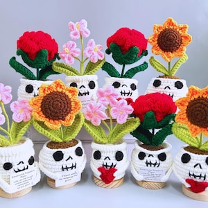 Puede incluir: Flores de ganchillo en macetas con forma de calavera. Las flores incluyen rosas rojas, flores rosas y girasoles. Las macetas son blancas con rasgos faciales negros. Algunas macetas tienen pequeñas etiquetas con texto.