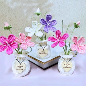May include: Handmade crochet flower arrangements in small white vases. The flowers are in shades of pink, purple, and white. Each vase has a wooden heart tag with the words "You Are My Sunshine" or "Smile Everyday".
