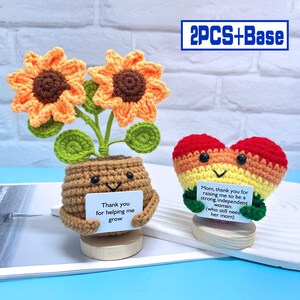 Può includere: Due figure all'uncinetto, una pianta di girasole e un cuore, ciascuna con un biglietto con un messaggio. La pianta di girasole ha un biglietto che dice "Grazie per avermi aiutato a crescere". Il cuore ha un biglietto che dice "Mamma, grazie per avermi cresciuta per essere una donna forte e indipendente. (che ha ancora bisogno della sua mamma)"