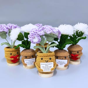 May include: A collection of handmade crochet plants in small pots. The plants feature white and purple flowers with green leaves. The pots are tan and yellow with smiling faces and hold signs that read "Emotional Support Plant".