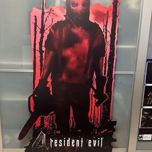 May include: A red and black cardboard cutout of a character from the video game Resident Evil 2. The character is holding a chainsaw and wearing a red shirt and a mask. The text "Resident Evil" and "PlayStation 2" are visible at the bottom of the cutout.