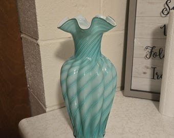 May include: A glass vase featuring a light turquoise and white swirled pattern with a vertical ribbed texture and a ruffled, flared rim. The body of the vase tapers toward the neck and widens at the base, showcasing a translucent, layered design.