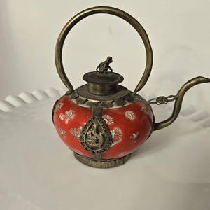 May include: A small, ornate teapot with a red and white floral design. The teapot has a brass handle, spout, and accents, including a small monkey figure on the lid. The teapot is likely made of porcelain and metal.