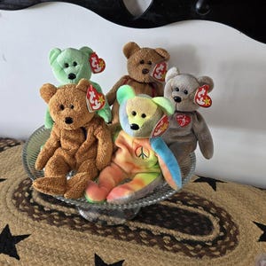 May include: Five colorful beanie baby plush toys, including a green bear, a brown bear, a brown bear with a peace sign, a gray bear, and a tie-dye bear, are displayed on a glass cake stand. The beanie babies are all sitting on a brown and black rug with stars.