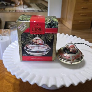 May include: A Hallmark Keepsake Starship Christmas ornament featuring a silver UFO with a clear dome containing Santa Claus and a reindeer. The ornament has the text Starship Christmas printed on the side and includes magic blinking lights.