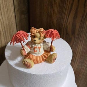 May include: A two-tiered white cake dummy with a small teddy bear figurine wearing an orange and white cheerleader outfit and holding two orange pom-poms. The teddy bear has a brown nose and brown eyes and is wearing a white shirt with the letters "CT" on it.