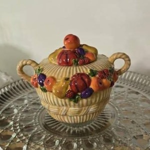 May include: A ceramic sugar bowl with a basket weave design, adorned with colorful fruit. The lid features a large orange fruit as a handle. The bowl has two handles and sits on a decorative glass tray.