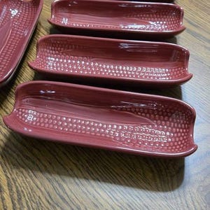 May include: A set of six burgundy-colored ceramic corn-shaped serving dishes. Each dish has a textured, dotted pattern in the center and curved edges. The dishes are arranged on a wooden table.