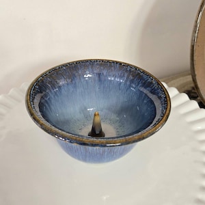 May include: A small, blue ceramic bowl with a raised center, likely for incense. The bowl has a gradient of blue hues and a brown rim. It sits on a white, ruffled plate, suggesting a decorative or functional item.