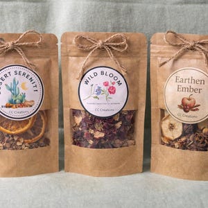 Puede incluir: Tres bolsas de papel marrón con ventanas transparentes, atadas con lazos de cuerda. Cada bolsa tiene una etiqueta circular con un diseño botánico y texto. Las bolsas contienen hierbas secas y rodajas de fruta. Las etiquetas dicen "Desert Serenity", "Wild Bloom" y "Earthen Ember".