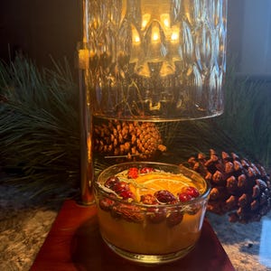 Può includere: Un scaldacandele decorativo con una ciotola di vetro piena di fette d'arancia, mirtilli rossi e spezie. Lo scaldacandele ha una cornice in metallo color oro e una base in legno. Pigne e verde sullo sfondo.