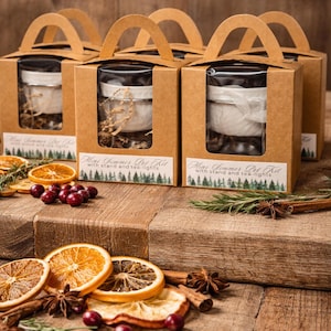 May include: Several gift boxes with handles, each containing a glass jar with a white candle and decorative elements. The boxes are brown with a clear window. Dried orange slices, cranberries, and spices are arranged on a wooden surface.
