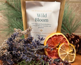 May include: A package labeled "Wild Bloom" from The Botanical Collection, with dried lavender, rosemary, and citrus slices on a wooden board. The package lists notes of wild rose, lavender, and geranium. A pine cone is also visible.