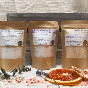 May include: Three brown kraft paper pouches containing bath salts, labeled Rejuvenating Rosemary and Eucalyptus, Relaxing Lavender, and Hello Sunshine. Each pouch features a clear window showing the salt contents, with dried botanicals and citrus slices.