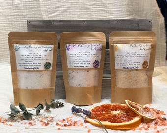 May include: Three brown kraft paper pouches containing bath salts, labeled Rejuvenating Rosemary and Eucalyptus, Relaxing Lavender, and Hello Sunshine. Each pouch features a clear window showing the salt contents, with dried botanicals and citrus slices.
