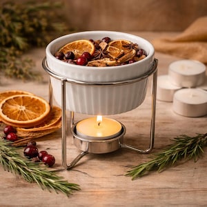 May include: A white ceramic bowl filled with dried orange slices, berries, and spices, set on a silver metal stand. A lit candle sits below the bowl, with additional orange slices and greenery nearby. The arrangement is on a wooden surface.