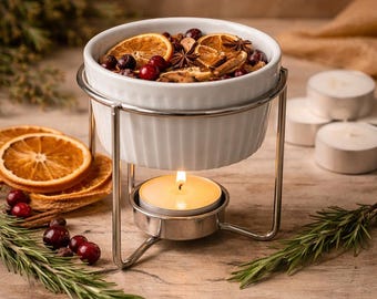 May include: A white ceramic bowl filled with dried orange slices, berries, and spices, set on a silver metal stand. A lit candle sits below the bowl, with additional orange slices and greenery nearby. The arrangement is on a wooden surface.