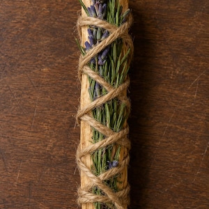 May include: A cylindrical piece of light brown wood wrapped in tan twine, securing a bundle of dried purple lavender sprigs and green rosemary needles against the surface of the wood, creating a textured, botanical, and rustic decorative item.