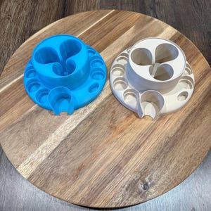 May include: Two 3D-printed objects, one light blue and one off-white, sit on a wooden surface. Each has a circular base with small holes and a central, heart-shaped design. A curved, U-shaped feature extends from the base.