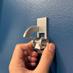 May include: A silver-colored, mechanical device with a curved, numbered scale and a rotating component. The device is mounted on a rectangular base and held against a blue wall.
