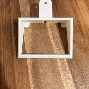 May include: A white, 3D-printed square-shaped object with a handle and a hole at the top. The object has a rectangular opening in the center and small supports on the sides. It is designed to be a cookie cutter.