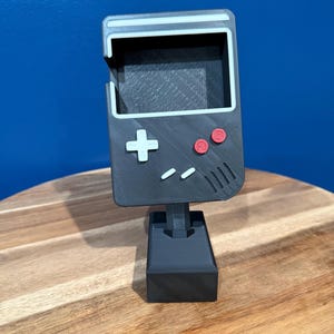 May include: A black and gray 3D-printed Game Boy-style phone stand. The stand features a screen, directional pad, and buttons in red and white. The stand is 6 inches tall and sits on a rectangular base.