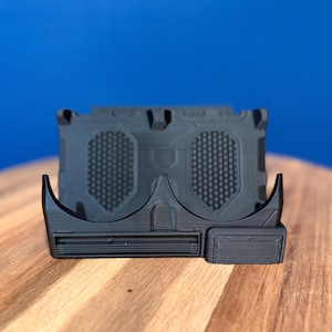 May include: A black 3D-printed device with a geometric design. The device has a curved front, a rectangular base, and a textured back panel with a grid pattern. The object is on a wooden surface with a blue background.