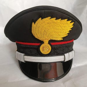 Puede incluir: Una gorra militar de plato negra con un prominente emblema bordado en amarillo de una granada en llamas en la parte frontal. La gorra incluye una fina banda horizontal roja, una banda negra texturizada, una correa plateada y una visera negra brillante.