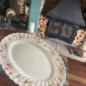 May include: A white oval serving platter with a floral design in shades of pink, yellow, and brown. The platter has a scalloped edge and is sitting on a wooden table.