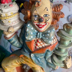 Puede incluir: Figura de payaso vintage con cara pintada, pelo rojo y atuendo azul y naranja. El payaso sostiene un pastel en capas y una pila de tazas de té. La figura tiene una estética caprichosa y antigua.