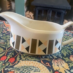 May include: Cream-colored ceramic gravy boat with a geometric pattern of brown, black, and peach triangles and rectangles. The gravy boat has a spout and a square handle. The gravy boat is on a floral patterned tablecloth.