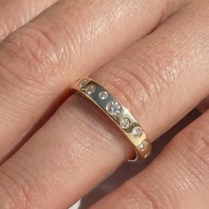 May include: A gold band ring with multiple clear gemstones set around the band. The ring is on a person's finger, with the hand and nails in the frame. The ring is a simple, elegant design.