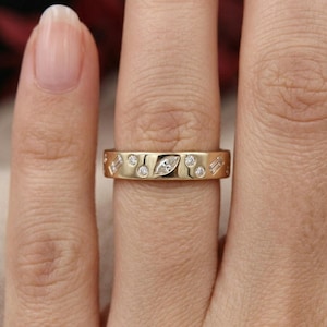 May include: A gold band ring with a series of small, round, and marquise-shaped diamonds. The ring is worn on a finger, showcasing the intricate design and sparkle of the gemstones. The ring's warm gold color contrasts with the skin tone.