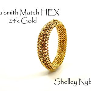 May include: A 24k gold ring with a textured, beaded surface pattern. The text Metalsmith Match HEX 24k Gold and Shelley Nybakke is visible in the image. The ring has a circular shape and a metallic yellow finish, displaying a repeating hexagonal bead design.