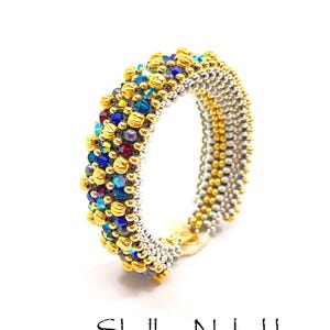 Könnte beinhalten: Ein handgefertigtes Perlenarmband mit einer Mischung aus goldenen, silbernen und farbigen Perlen. Das Armband hat ein zentrales Band aus silbernen Perlen, akzentuiert mit Gold und einer Ansammlung von blauen, roten und irisierenden Perlen. Der Name des Designers, Shelley Nybakke, steht unten.