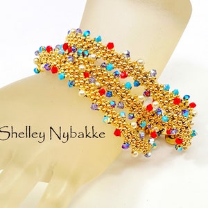 Könnte beinhalten: Ein goldfarbenes Armband mit roten, blauen und weißen Perlen. Das Armband befindet sich an einer Mannequinhand.