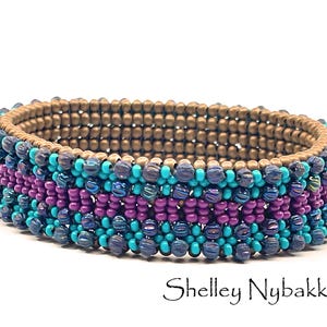 Op de afbeelding: Een kralenarmband met rijen van metallic bronskleurige, levendig turquoise, dieppaarse en iriserende donkerblauwe kralen. De armband heeft een gestructureerd, meerlaags ontwerp met een consistent patroon. De tekst Shelley Nybakke is zichtbaar.