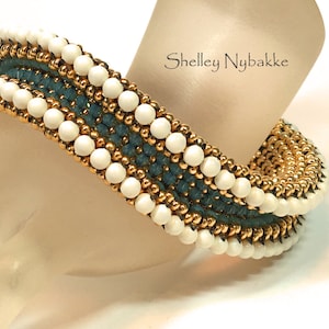 Puede incluir: Un brazalete de cuentas doradas, blancas y azules con un diseño tejido. El brazalete está en el brazo de un maniquí. Shelley Nybakke está impreso en el brazo del maniquí.