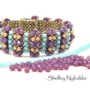 May include: A beaded bracelet featuring alternating vertical rows of faceted purple crystals, light blue beads, and metallic gold spheres. Loose light blue and purple beads are scattered in the foreground. The text Shelley Nybakke is visible.