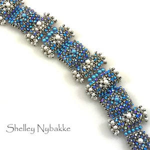 Könnte beinhalten: Ein Perlenarmband mit einem Design aus blauen, weißen und irisierenden Perlen. Das Armband hat einen silberfarbenen Verschluss und besteht aus kleinen, runden Perlen. Der Name "Shelley Nybakke" ist in Schreibschrift geschrieben.