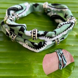 May include: A fabric bracelet with a green, black, and white pattern. The bracelet is adorned with silver-colored metal rings. The image also shows a wrist wearing the bracelet.