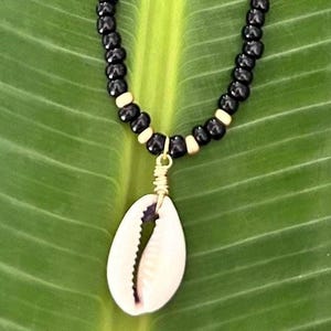May include: A necklace featuring a cowrie shell pendant. The necklace has a beaded design with black and tan beads. The pendant is a cream color and is attached with gold-colored wire. The necklace is displayed on a green leaf.